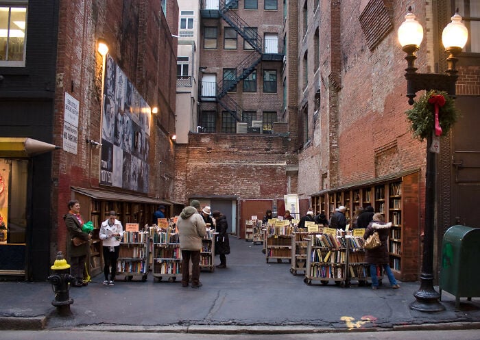 Gem&uuml;tlicher Buchladen im Freien in einer urbanen Gasse mit Menschen, die B&uuml;cher durchst&ouml;bern, umgeben von Ziegelmauern und Vintage-Stra&szlig;enlaternen.