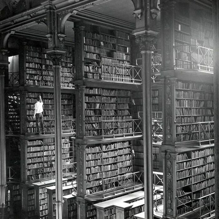 Gro&szlig;artiges Bibliotheksinterieur mit hohen B&uuml;cherregalen, mehreren Balkonen und einer Person, die in einer magischen Umgebung nach einem Buch greift.
