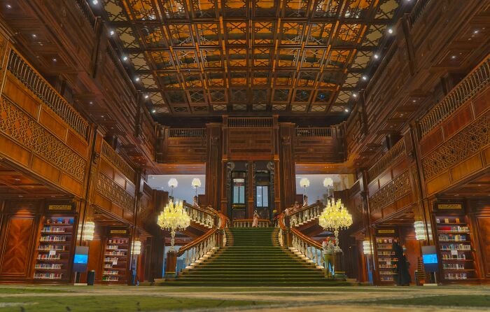 Grandiose Bibliothekseinrichtung mit einer gro&szlig;en Treppe, h&ouml;lzernen verzierten W&auml;nden und Regalen voller B&uuml;cher in einer magischen Umgebung.