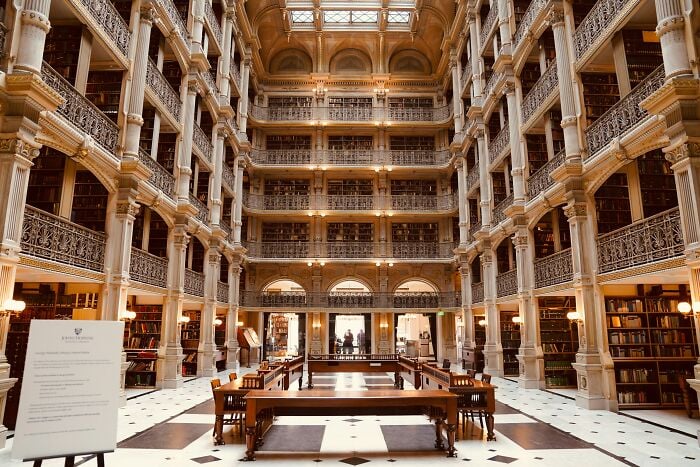 Grandiose Bibliothekseinrichtung mit mehreren Ebenen von Bücherregalen, verzierten Säulen und einem geräumigen Lesebereich mit Holztischen und Stühlen. Grandiose Bibliothekseinrichtung mit mehreren Ebenen von Bücherregalen, verzierten Säulen und einem geräumigen Lesebereich mit Holztischen und Stühlen.