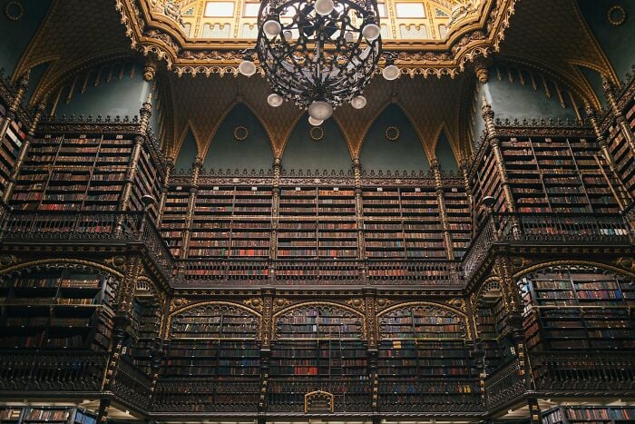 Pr&auml;chtiger, grandioser Bibliotheksinnenraum mit hoch aufragenden B&uuml;cherregalen und filigranen Golddetails unter einem dekorativen Kronleuchter.