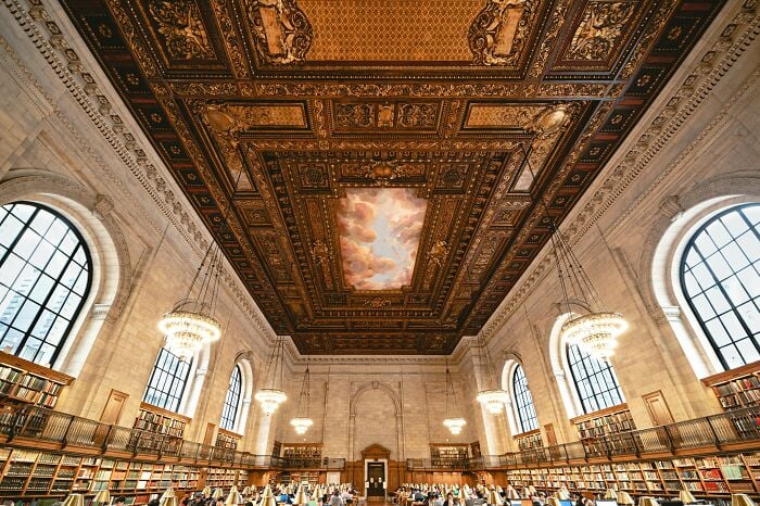 Grandiose Bibliothekseinrichtung mit kunstvoll verzierter Decke, gro&szlig;en Bogenfenstern, Kronleuchtern und Reihen von B&uuml;cherregalen voller B&uuml;cher.